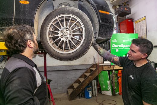 Two Auto Solve mechanics inspecting a vehicle's wheel and suspension on the lift during a winter safety check